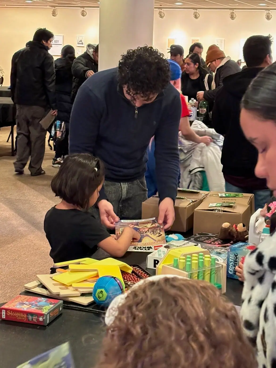 A volunteer helping a young girl pick a book at the toy table.